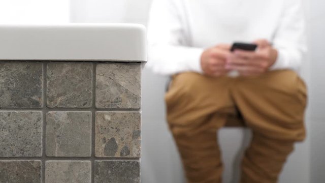 A man with trousers down sitting on the toilet and using a smartphone.