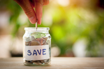 Hand putting coins in a jar. Saving money concept and save money to support everything in life with sun light bokeh background.On wooden table.