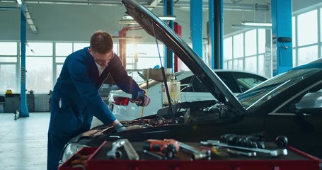 Male Caucasian young auto mechanic standing at open car and repairing motor in big garage. Man in uniform and goggles servicing vehicle in automobile repairment salon indoor. Transport service concept - Powered by Adobe