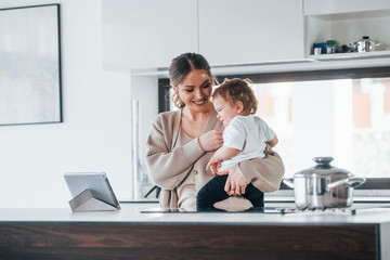 Female blogger mother with her kid together indoors at home at weekend using tablet