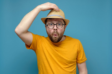 Surprised Caucasian man in round glasses clings to his hat, as if greeting someone, on a blue background.