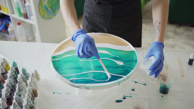 Tracking from above shot of female artist in protective gloves using wooden stick to pour white acrylic paint mixed with epoxy resin onto turquoise color on round canvas to make fluid painting