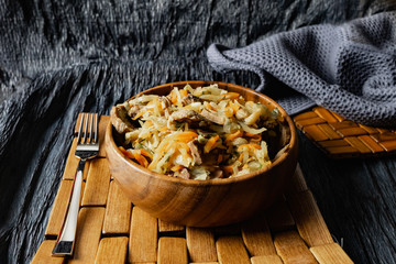 Stewed cabbage with oyster mushrooms and pork tongue with carrots and onions. Hot dish in a wooden plate on a wooden stand, next to a fork, a sprig of grass and a napkin. Black background with relief
