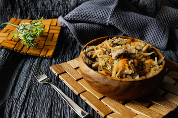 Stewed cabbage with oyster mushrooms and pork tongue with carrots and onions. Hot dish in a wooden plate on a wooden stand, next to a fork, a sprig of grass and a napkin. Black background with relief