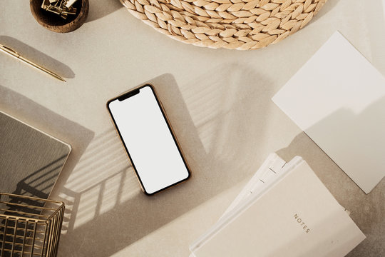 Flatlay of blank screen smart phone, notebooks, clips in wooden bowl, straw stand on beige concrete background. Home office desk workspace. Business, work template. Flat lay, top view.