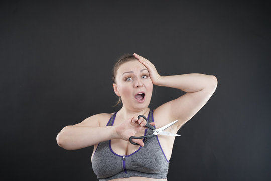 Plus Size Woman Shows Her Hairy Armpit. The Girl Holds A Pair Of Scissors In Her Hand And Experiences Negative Surprised Emotions, Hating Excessive Body Hair . Studio Black Monochrome Background.