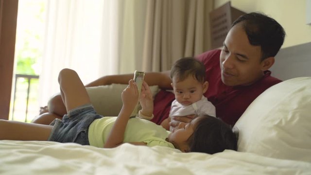 asian father and his two daughters lay in bed while looking at the video with a smartphone