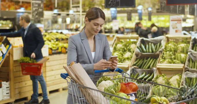 Smiled beautiful Caucasian woman leaning on trolley full of food in supermarket and tapping or texting message on smartphone. Female customer typing and scrolling on phone with shopping list.