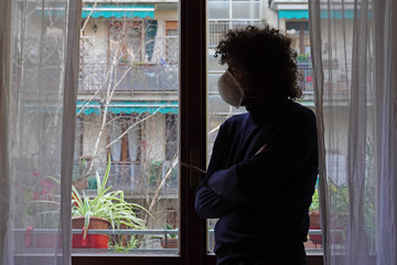 Europe, Italy, Milan - Man 40 years old at home in quarantine with mask looks out of the closed window to protect himself from the pandemic of n-cov19 Coronavirus  