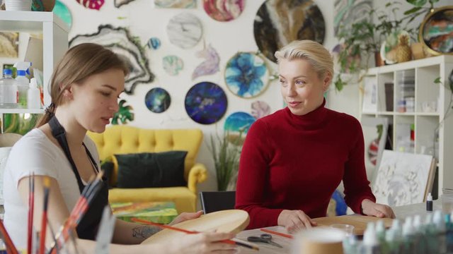 Medium shot of art teacher explaining middle aged female student how to paint on round wooden canvas sitting at desk in art studio