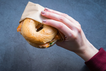 Woman's hand holding a bagel filled with salmon, avocado, cream cheese and arugula on concrete background.