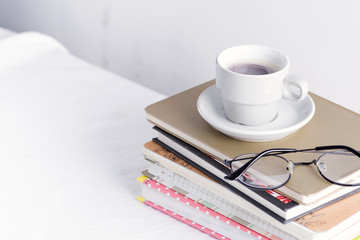 Stack of different books and notepads with woman's glasses and coffee cup on the top on a white table.