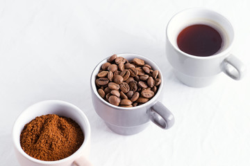 Three cups of different condition of coffee - beans, freshly ground and coffee drink on a marble light grey background.