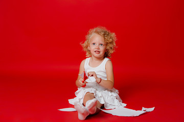 Cute blonde curly girl cutting paper with red scissors isolated on red