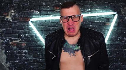 Handsome young man with glasses in leather jacket on naked body with tattoo on chest shouting and waving his hands on background brick wall with glowing lamps. Angry male expressing negative emotions. - Powered by Adobe