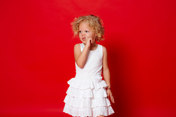 Pretty little blonde curly girl in white dress touching her nose isolated on red