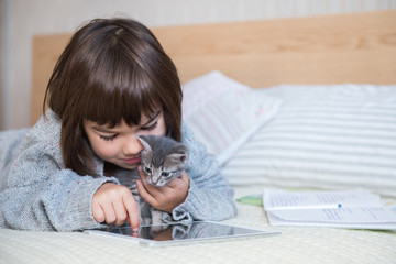 cute girl schoolgirl 7 years old does lessons on a tablet distance learning holds gray kitten in bed