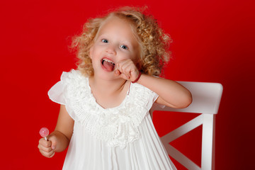 Beautiful little curly blonde girl eating lollipop sitting on white chair isolated on red