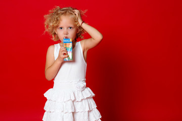 Cute curly girl in white dress drinking juice isolated on red background