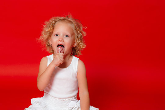 Beautiful little curly blonde girl eating lollipop sitting on the floor isolated on red