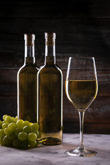 Two full white wine bottle and wineglass with grapes on a white cloth on a wooden background