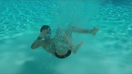 Boy in swimming goggles is diving underwater in the pool and waving his hand at the camera
