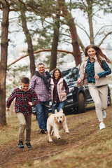 Happy family runs and having fun with their dog near modern car outdoors in forest