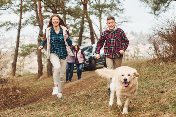 Happy family runs and having fun with their dog near modern car outdoors in forest