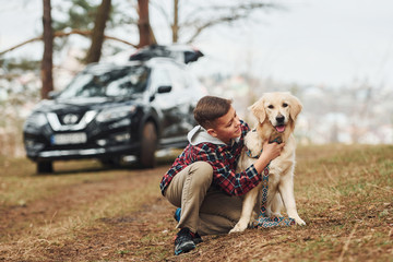 Cheerful boy in casual clothes sitting with her dog in forest against modern black car