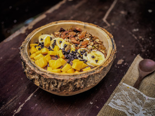 close-up of a vegan fruit smoothie bowl with wooden spoon, granola, nuts, banana mango and cocoa on a table for a healthy breakfast 