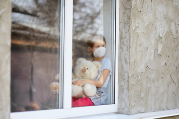 Sad Child, little girl in mask with teddy bear looking from window, coronavirus quarantine