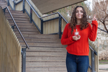 Portrait of attractive caucasian young woman with cologne perfume bottle keeping finger crossed, having calm expression, in the park, orange sweater and jeans, long curly hair. Place for your text in