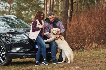 Man with woman have fun with their dog outdoors in forest near modern black car