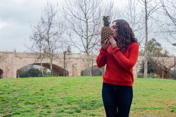 Portrait of healthy attractive caucasian young woman kissing pineapple. Dieting concept. Healthy lifestyle with fruit, in the park, orange sweater and jeans, long curly hair. Place for your text in co