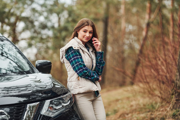 Fototapeta na wymiar Young cheerful girl standing near modern black car outdoors in the forest
