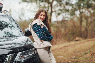Young cheerful girl standing near modern black car outdoors in the forest