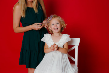 Mother curls daughter hair. Little blonde girl sitting on a white chair isolated on red
