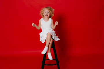 Amuzing funny little blonde girl in white dress sitting on chair isolated on red