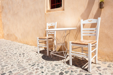 Fototapeta na wymiar Two white wooden chairs with table on the street. Traditional greek architecture on Santorini island, Greece.
