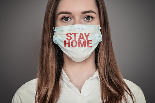 Portrait of a scared young woman wearing a medical respiratory mask with stay home words isolated over dark grey background, coronovirus epidemic, quarantine, pandemic, isolation concept
