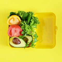 Lunchbox with a snack from lettuce, tomato, avacado on a yellow background. Healthy eating concept. Go vegan. Tasty vegetarian food in plastic box.