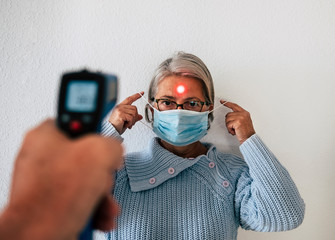 Temperature is checked for a senior woman - a retired person with a mask to avoid COVID-19 coronavirus infection. Prevention and cure concept
