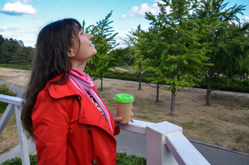 Outdoors portrait of cheerful beautiful brunette woman in red coat holding disposable coffee cup...