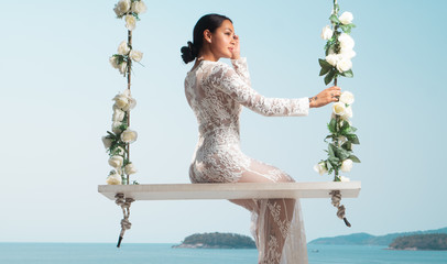 Beautiful woman in white lace dress posing on the outdoor swing with flowwers over sea and sky...