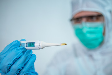 Closeup man wearing a protective suit and surgical mask, looking at thermometer