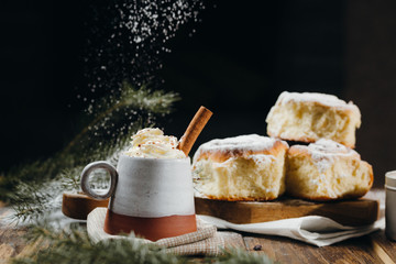 A hot cocoa with whipped cream, cinnamon stick and fresh Christmas buns with powder and over festive table, sugar powder is sprinkled on the background lke a snow. Winter holidays.