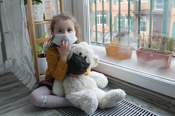 A girl and a toy bear in medical masks near the window, looking at the street. Schools and kindergartens are under quarantine. Concept of isolation and quarantine in the context of a pandemic. 