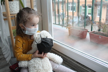 A girl and a toy bear in medical masks near the window, looking at the street. Schools and kindergartens are under quarantine. Concept of isolation and quarantine in the context of a pandemic. 