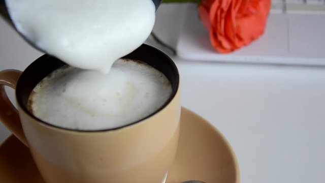 Pouring cream into a cup of coffee cappuccino whipped cream on a desktop next to a laptop and rose flower