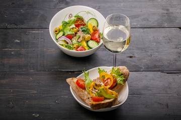 Food still life. Salad with vegetables and microgreens, canapes with pate, white wine in a glass on a dark wooden table.
